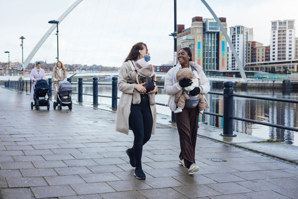 Group of young women walking together along a path with their babies in the city in Newcastle-upon-Tyne, North East England. They are wrapped up in warm clothing on a cold day, some mothers have their babies in a pram and some have their babies in baby harnesses.
