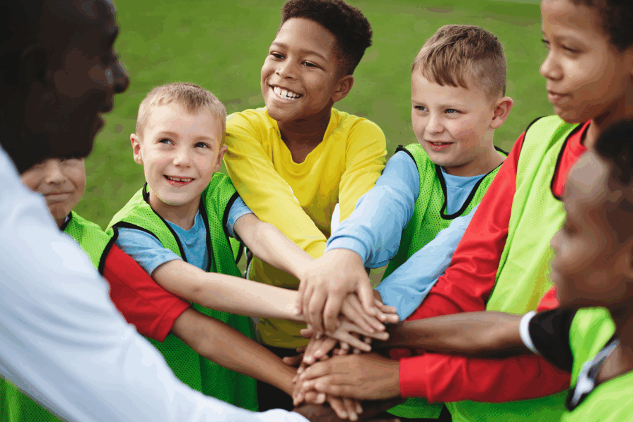 A group of children wearing sports bibs stand in a circle with their hands stacked together in the center.